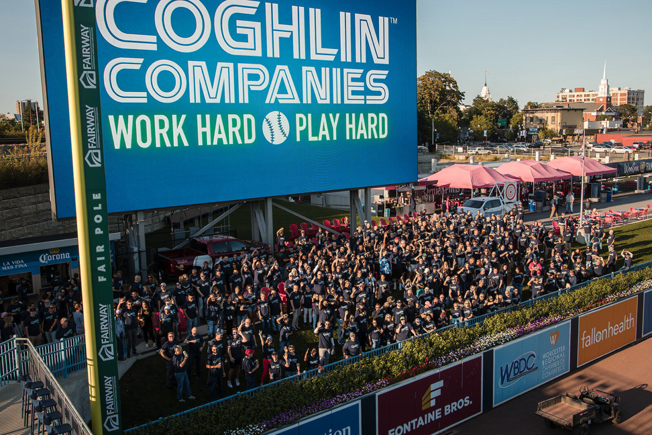 Coghlin Companies leadership team, Caring Associates, and family members come together at Polar Park for an employee celebration. The crowd of happy people stand under a large LED sign that reads 'Coghlin Companies: Work hard, play hard'